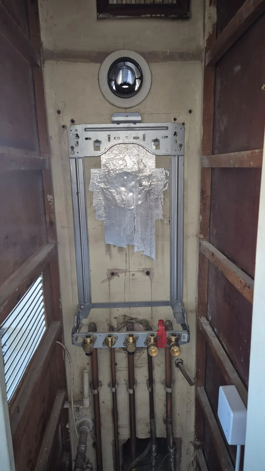 Old boiler out. Galvanised mounting frame fitted and a pre-plumbed manifold made up — brass isolation valves, red gas cock, five copper tails and the new flue elbow above.