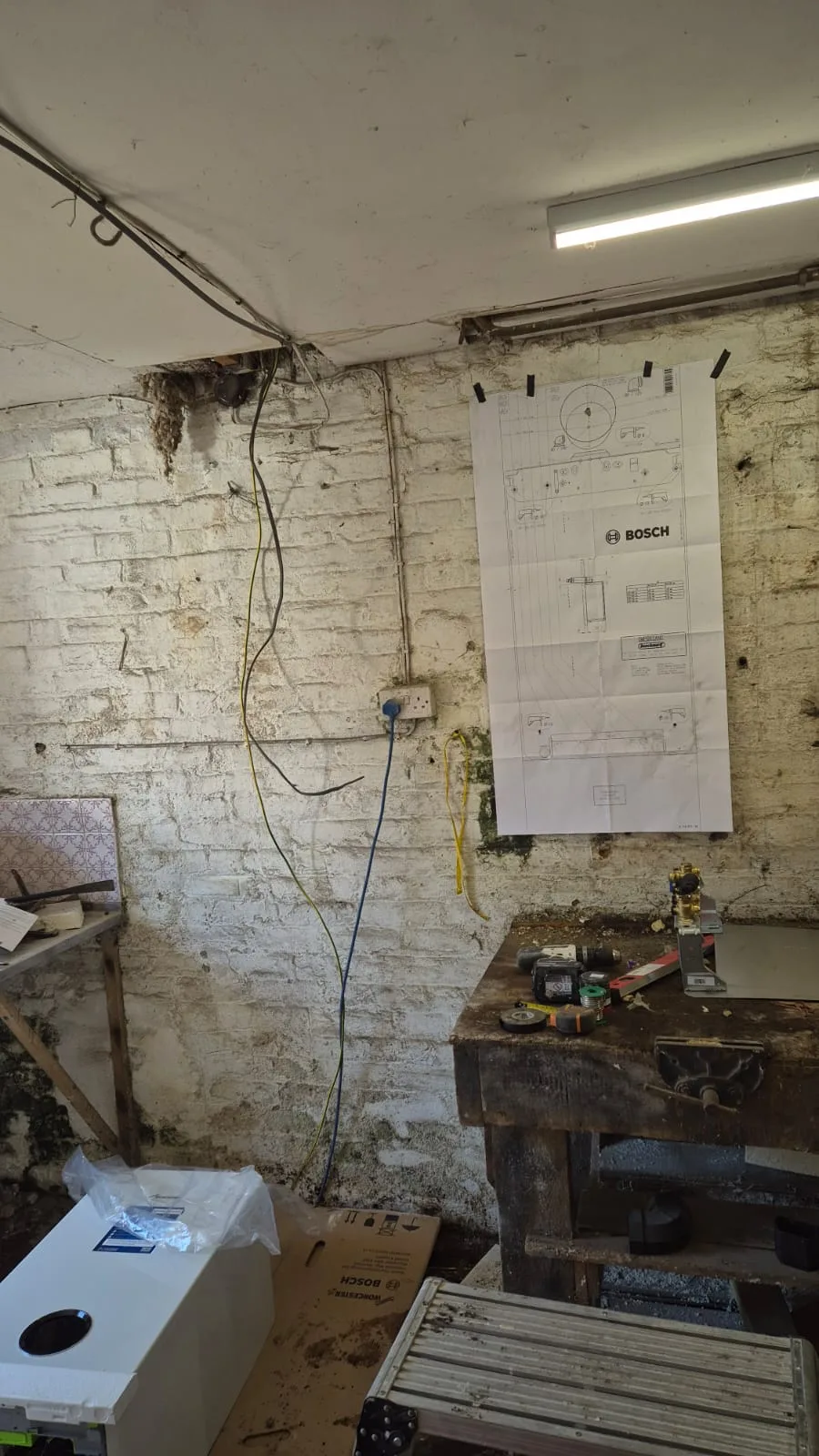 Bosch paper installation template taped to bare brick to mark pipe and flue positions before anything is drilled. The new boiler is still boxed on the floor.